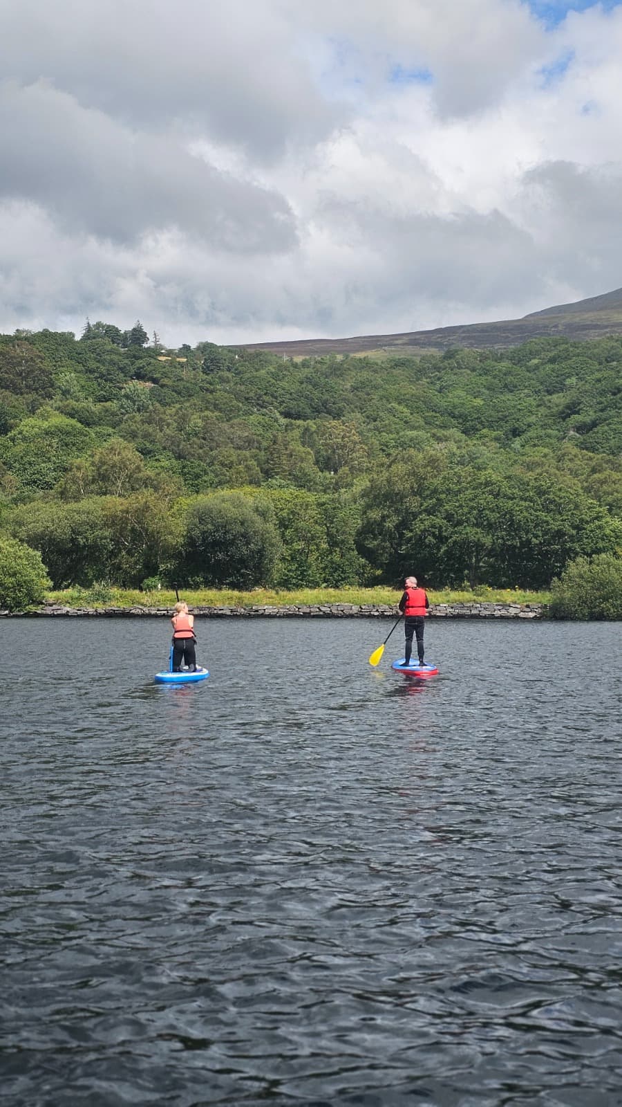 Two paddleboarders crossing a calm upland lake below wooded hills
